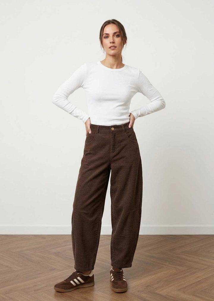 A woman stands in a studio wearing a fitted white long-sleeve top, dark brown wide-leg pants, and brown sneakers against a plain light wall and wood floor.
