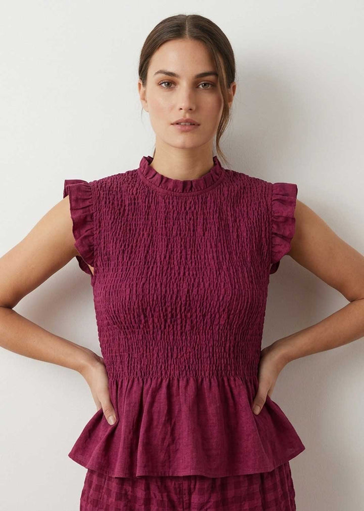 A woman modeling a sleeveless smocked peplum top in a deep berry purple color, paired with matching textured bottoms, against a plain off-white wall.