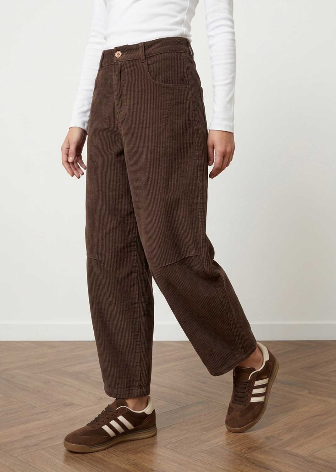 A person wearing dark brown corduroy wide-leg trousers, styled with a white long-sleeve top and brown Adidas sneakers, standing on a wooden floor against a white wall.