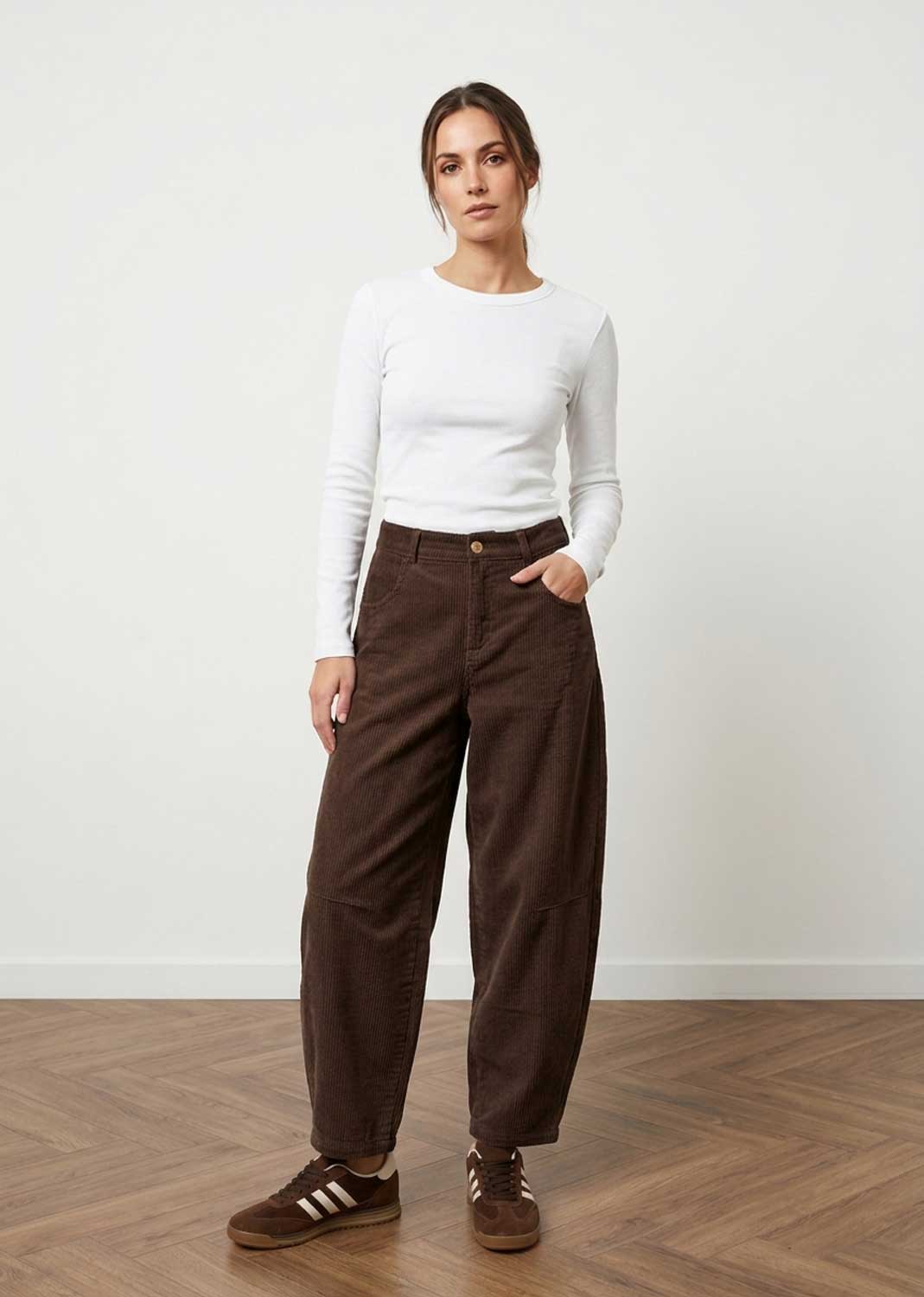 A woman modeling dark brown wide-leg corduroy pants, paired with a white long-sleeve top and brown sneakers, standing in a minimalist room with a light wall and wooden floor.