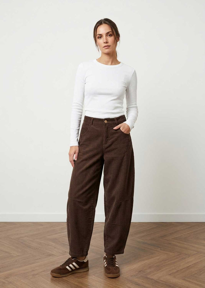 A woman modeling dark brown wide-leg corduroy pants, paired with a white long-sleeve top and brown sneakers, standing in a minimalist room with a light wall and wooden floor.