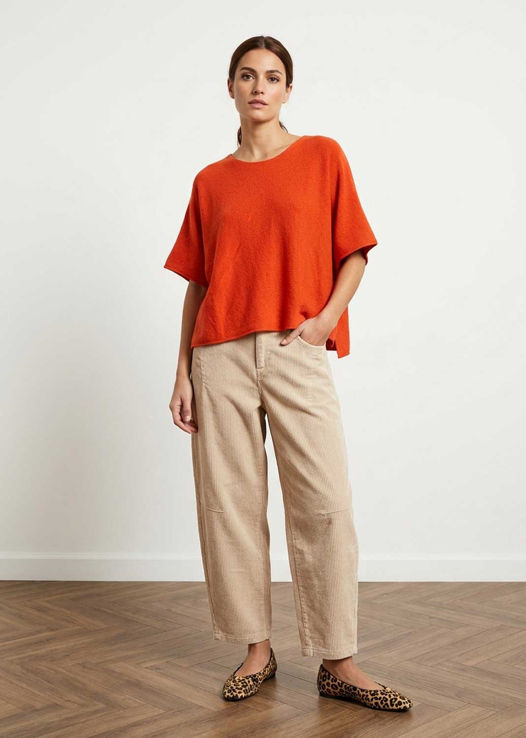 A woman stands in a studio wearing a loose bright orange short-sleeve top, beige wide-leg cropped pants, and leopard-print flats against a white wall and wood floor.