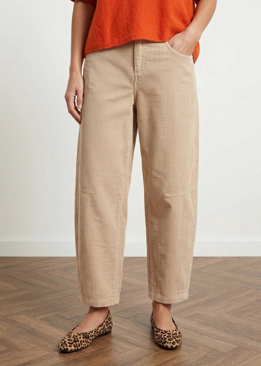 A person wearing beige corduroy cropped pants, an orange top, and leopard-print flats, standing on a wood floor against a white wall.