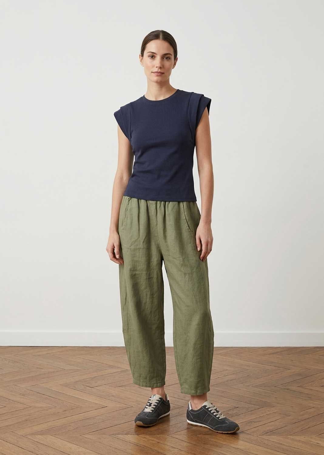 A woman stands in a studio wearing a navy blue sleeveless top and olive green wide-leg cropped pants with dark sneakers, against a plain light wall and wood floor.