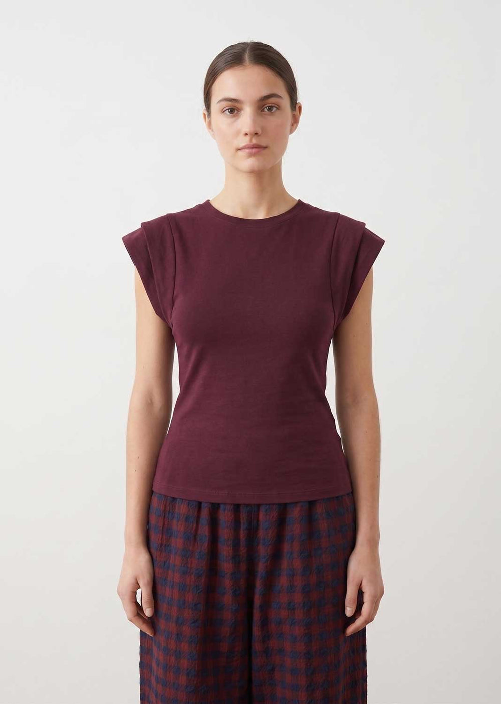 A woman modeling a short-sleeve crewneck top in deep burgundy, paired with dark blue and red checkered pants, against a plain light gray background.