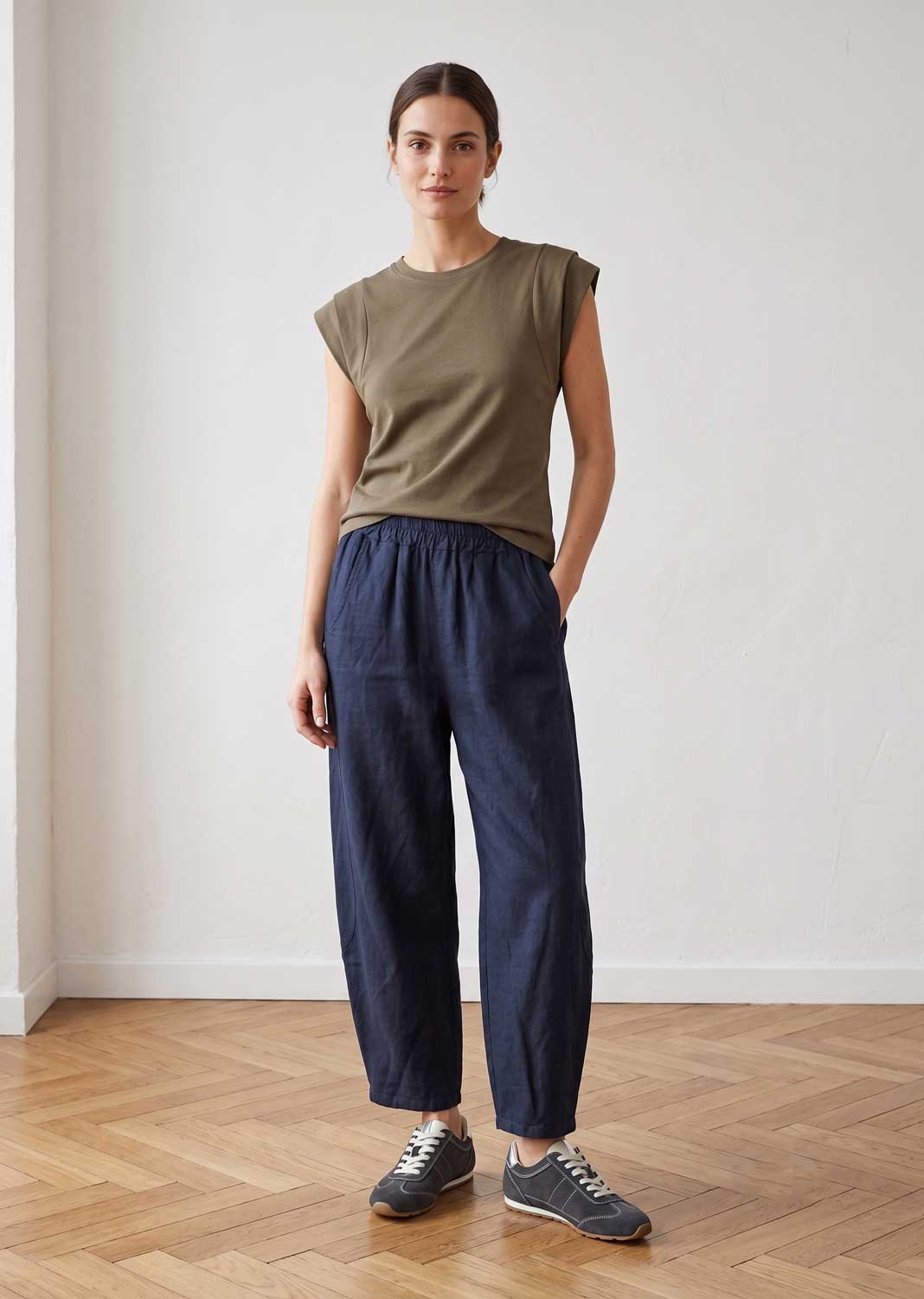 A woman stands in a bright indoor room wearing an olive green sleeveless top, loose dark navy pants, and gray sneakers.
