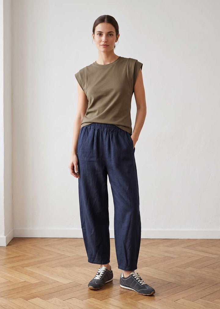 A woman stands in a bright indoor room wearing an olive green sleeveless top, loose dark navy pants, and gray sneakers.
