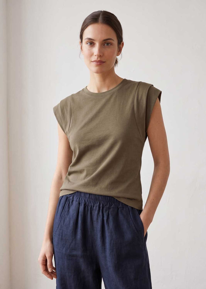A woman modeling a sleeveless olive green top with a crew neck and dark navy blue pants, standing against a plain off-white wall.