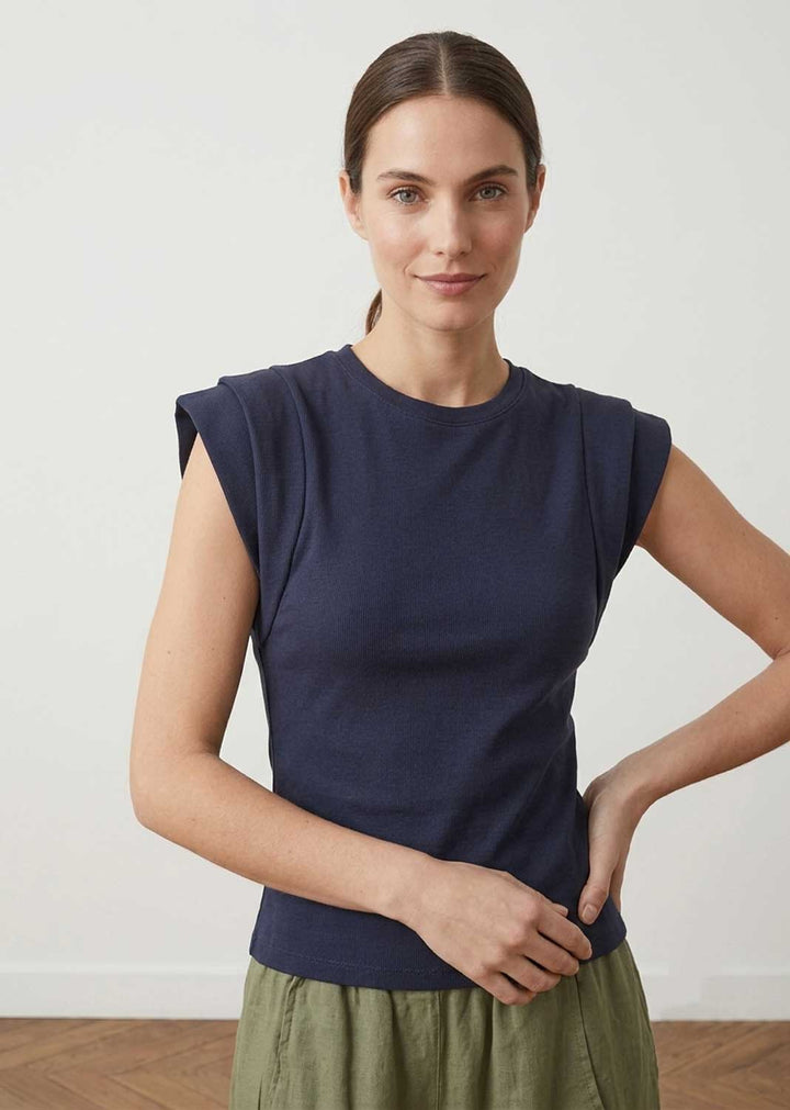 A woman modeling a sleeveless navy blue top with a relaxed, draped fit and an olive green skirt, standing against a plain off-white wall.