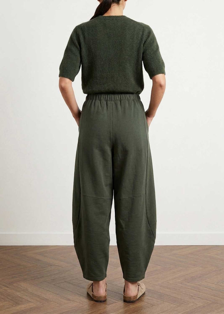 A person viewed from the back wearing an olive green short-sleeve knit top and matching wide-leg trousers, standing on a wood floor against a plain light wall.