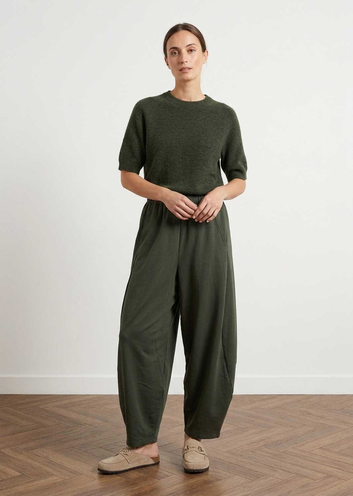 A woman stands in a minimalist room with a light gray wall and wood herringbone floor, wearing an olive green short-sleeve knit top and matching wide-leg trousers with beige slip-on shoes.