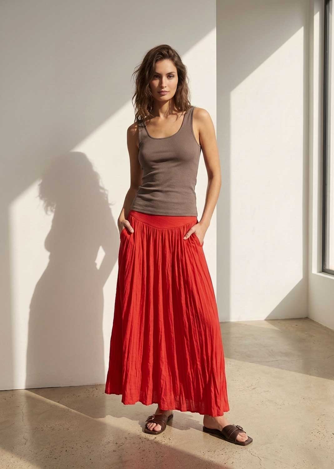 A woman stands in a bright room with strong sunlight and shadows, wearing a brown ribbed tank top and a long pleated red skirt, paired with dark brown slide sandals.
