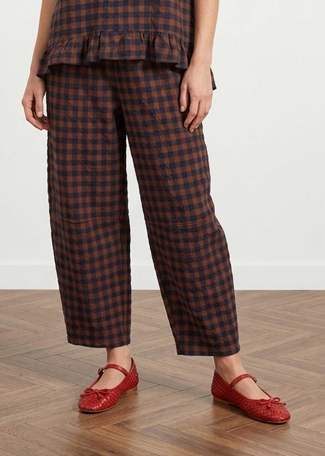 A person wearing loose dark brown and black checkered cropped trousers with a matching ruffled top and red Mary Jane flats, standing on a wood floor against a white wall.