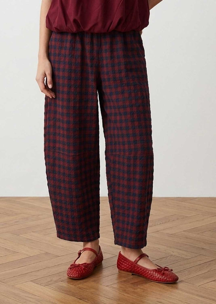 A person wearing loose cropped plaid pants in dark red and navy, paired with a burgundy top and red woven Mary Jane flats, standing on a light wood herringbone floor against a plain off-white wall.