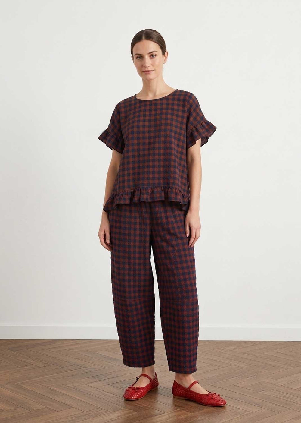 A woman modeling a matching two-piece plaid lounge set consisting of a short-sleeve top with ruffled edges and wide-leg cropped pants, in a dark red and black check pattern, paired with red flats.