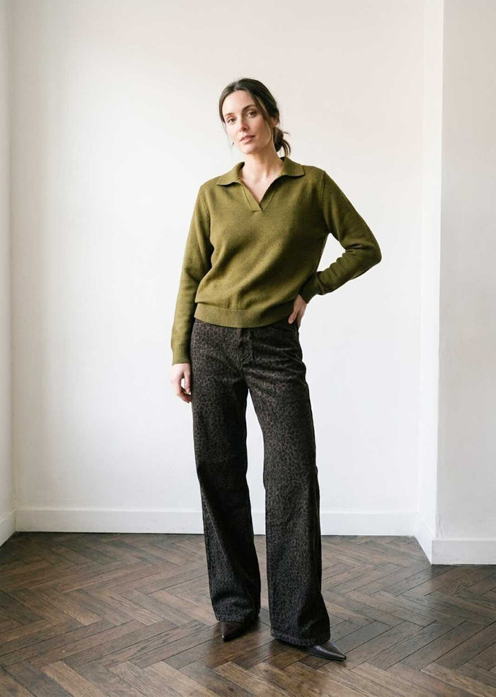 A woman stands in a minimalist room with white walls and a dark wood herringbone floor, wearing an olive green long-sleeve polo-neck sweater and dark patterned wide-leg trousers.