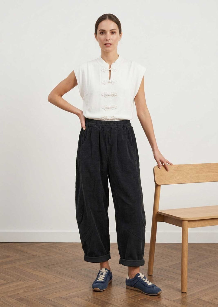 A woman stands in a studio wearing a sleeveless white top with front buttons and dark wide-leg pants, paired with blue sneakers. She is posed next to a light wood chair against a plain white wall and wood floor.
