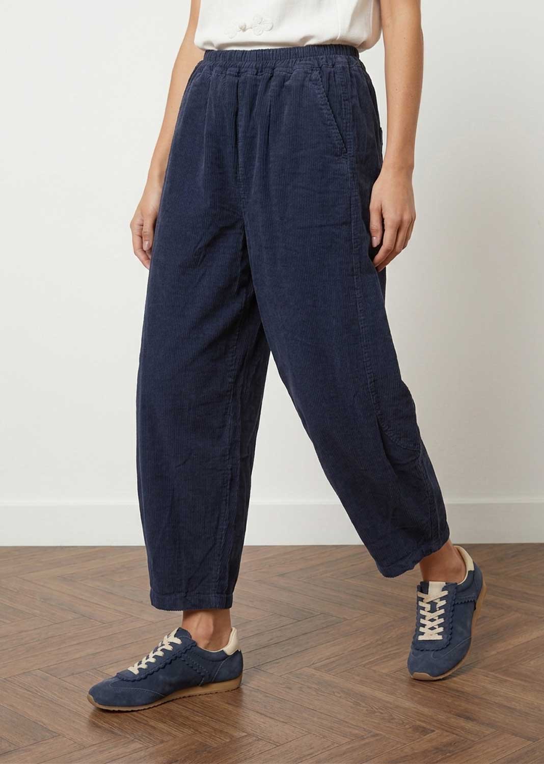 A person wearing loose dark navy wide-leg cropped barrel leg pants with an elastic waistband, paired with a white top and navy lace-up sneakers, standing on a wood floor against a plain light wall.