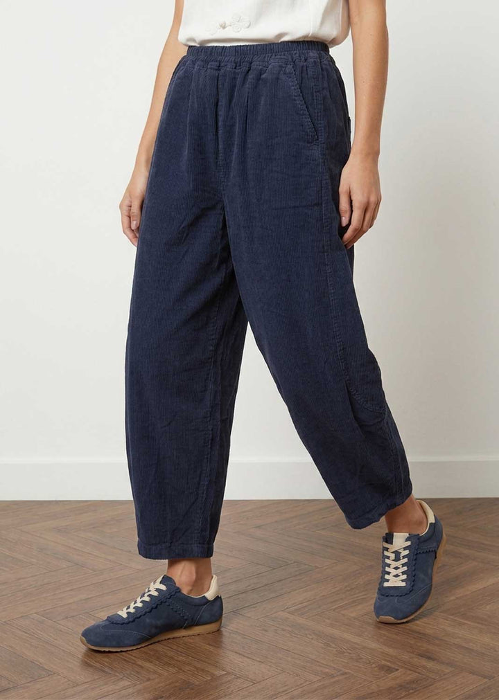 A person wearing loose dark navy wide-leg cropped barrel leg pants with an elastic waistband, paired with a white top and navy lace-up sneakers, standing on a wood floor against a plain light wall.