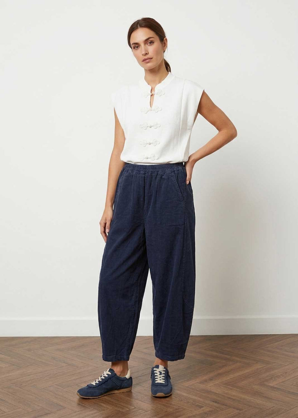 A woman modeling a white sleeveless button-front top and navy barrel leg cropped pants with blue sneakers, standing against a plain white wall on a wood floor.
