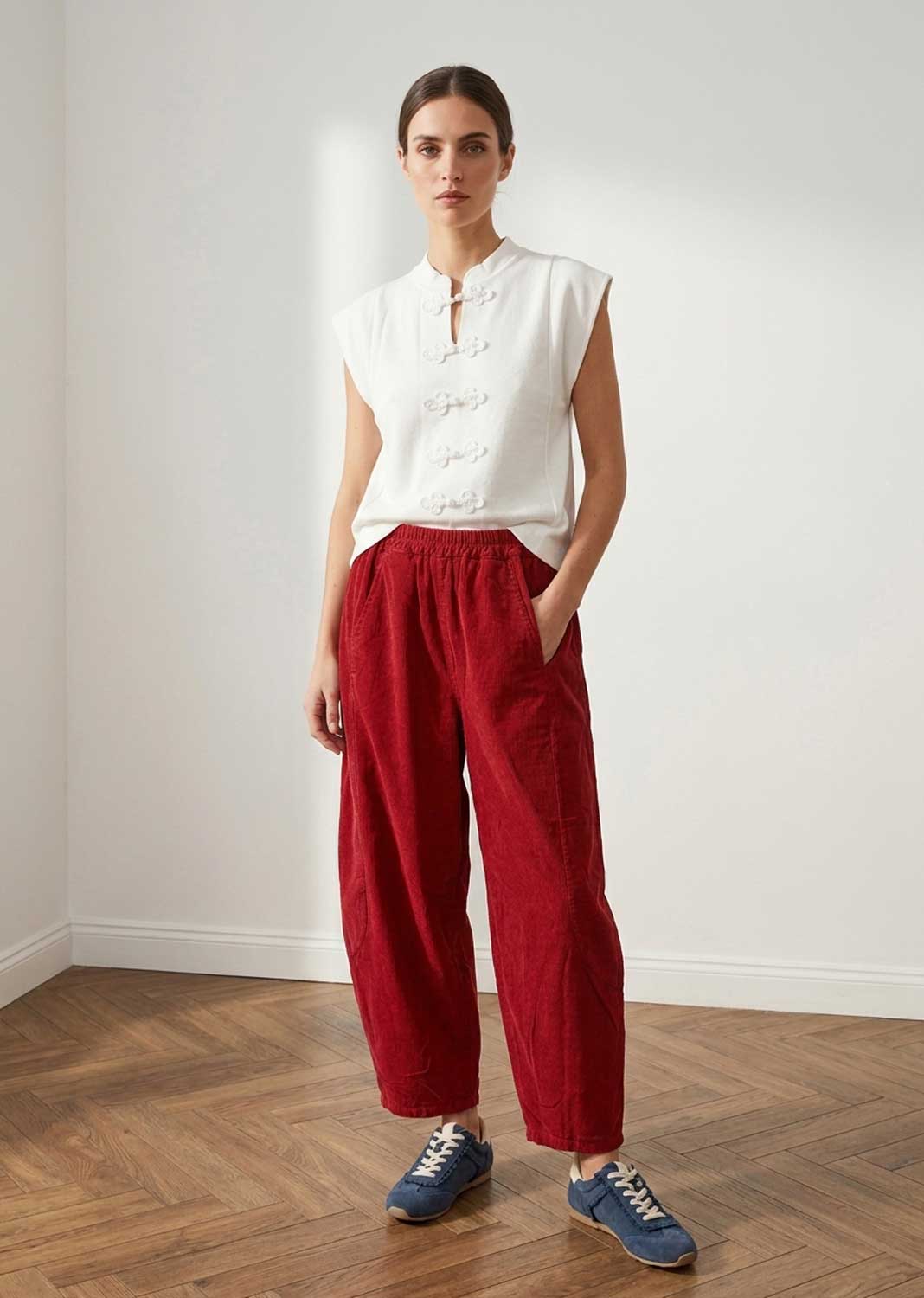 A woman stands in a minimalist room with a white wall and wood floor, wearing a white sleeveless top with decorative buttons and a high collar, paired with loose red barrel leg needlecord pants and blue sneakers.
