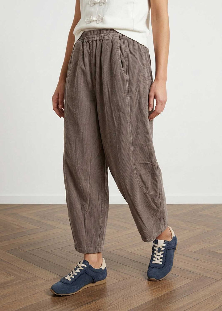 A person wearing loose, baggy barrell eg needlecord mocha pants with an elastic waistband and side pockets, paired with a white sleeveless top and blue sneakers, standing on a wooden floor against a plain white wall.