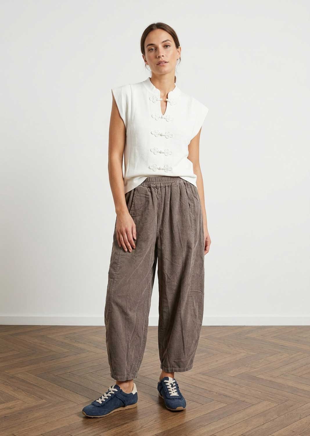 A woman stands in a studio wearing a sleeveless white top with decorative front buttons and loose taupe wide-leg barrel leg needlecord pants, paired with blue sneakers, against a light wall and wood floor.