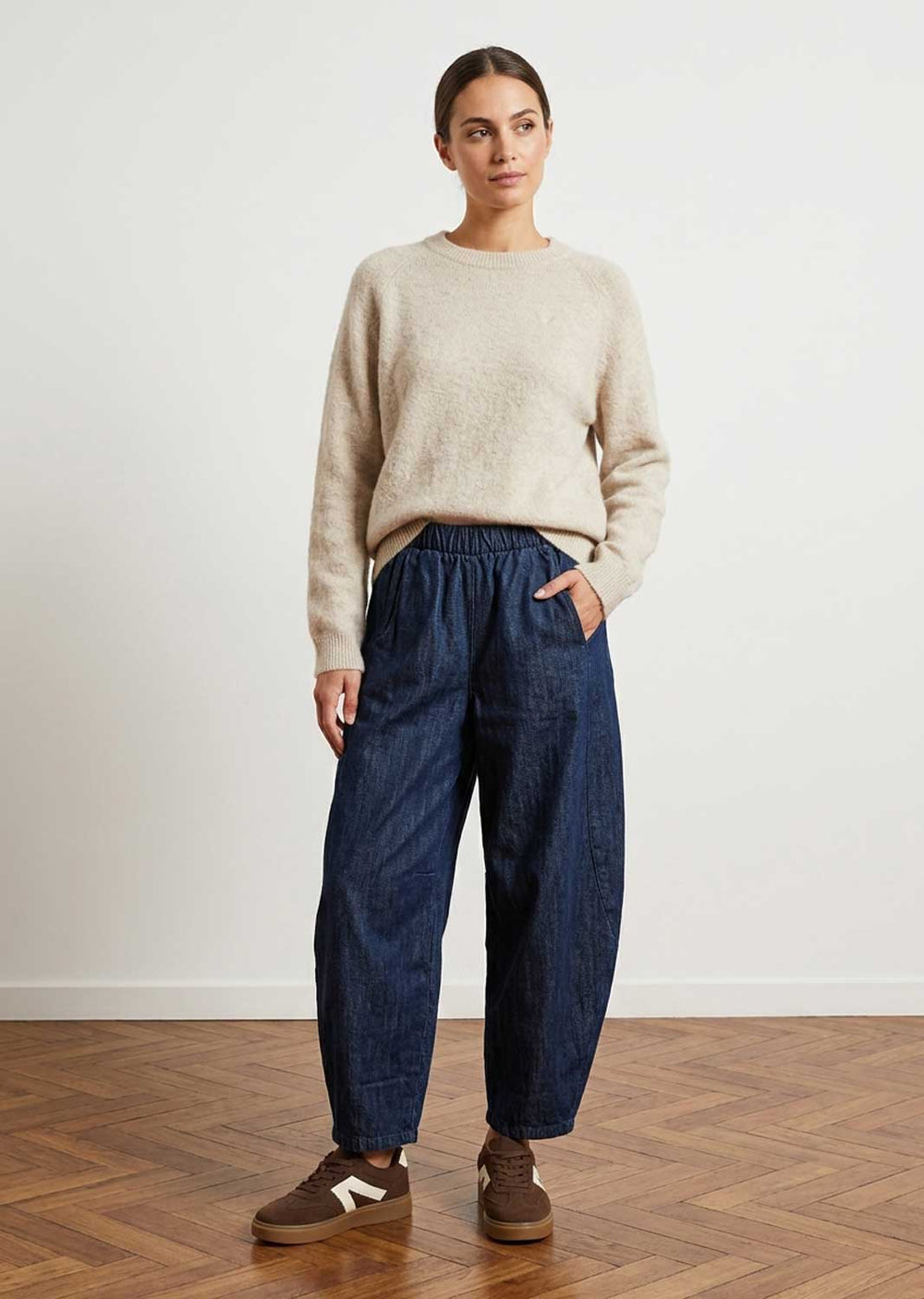 A woman stands in a studio wearing a fuzzy light beige crewneck sweater, dark blue barrel denim pants, and brown sneakers against a white wall and wood floor.