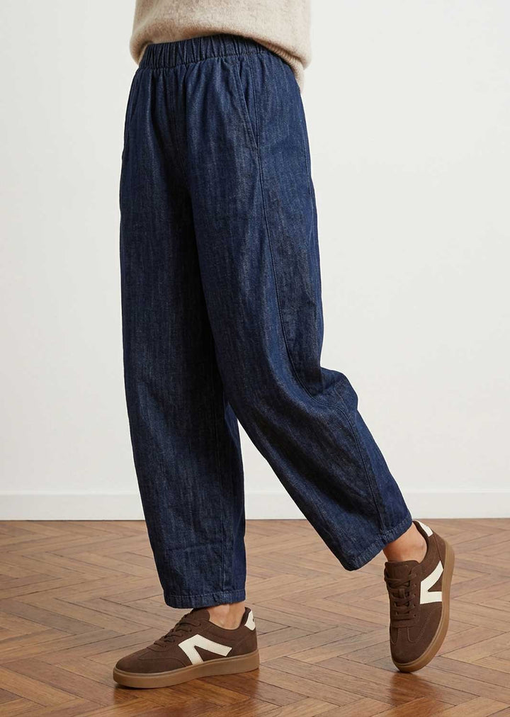 A person wearing loose dark blue denim pants and brown sneakers, shown from the waist down against a plain light wall and wood floor.