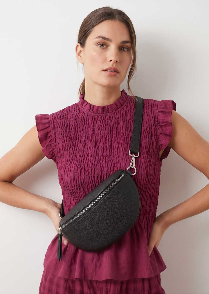 A woman wearing a textured magenta smocked dress models a black crescent-shaped crossbody bag with a silver-tone clasp and black strap against a plain white background.
