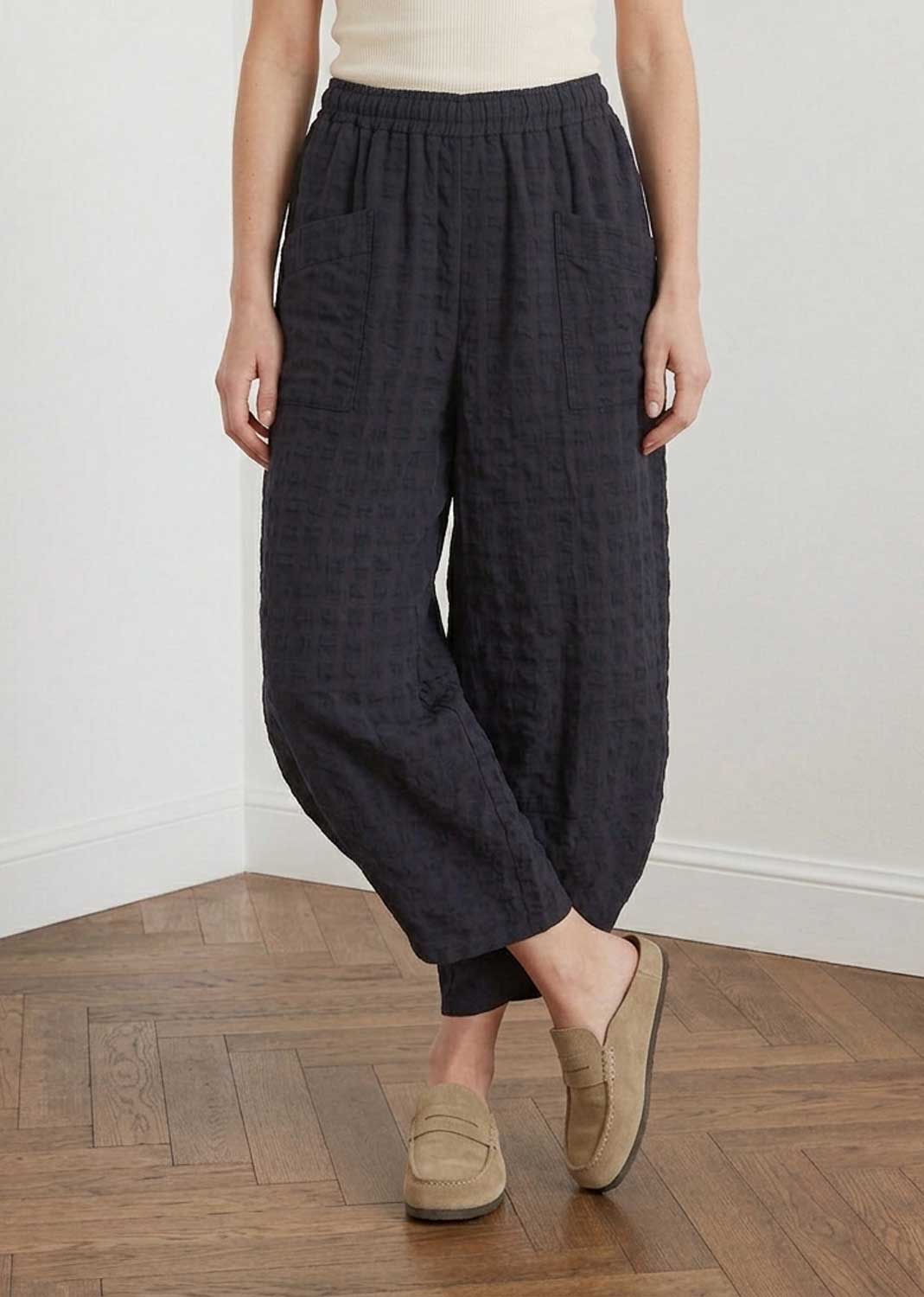 A person wearing loose, dark charcoal gray textured cotton harem-style pants with an elastic waistband, paired with beige slip-on loafers, standing on a wooden parquet floor against a white wall.