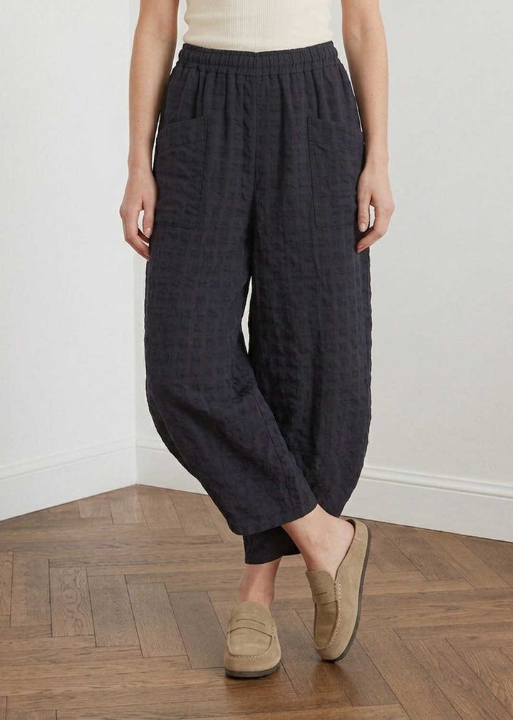 A person wearing loose, dark charcoal gray textured cotton harem-style pants with an elastic waistband, paired with beige slip-on loafers, standing on a wooden parquet floor against a white wall.
