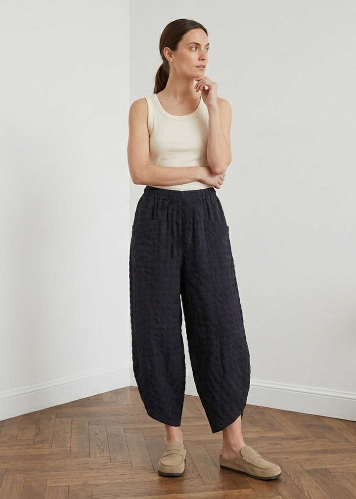 A woman stands in a minimalist room with a light gray wall and wood floor, wearing dark navy textured wide-leg cropped pants, a cream ribbed tank top, and beige slip-on shoes.