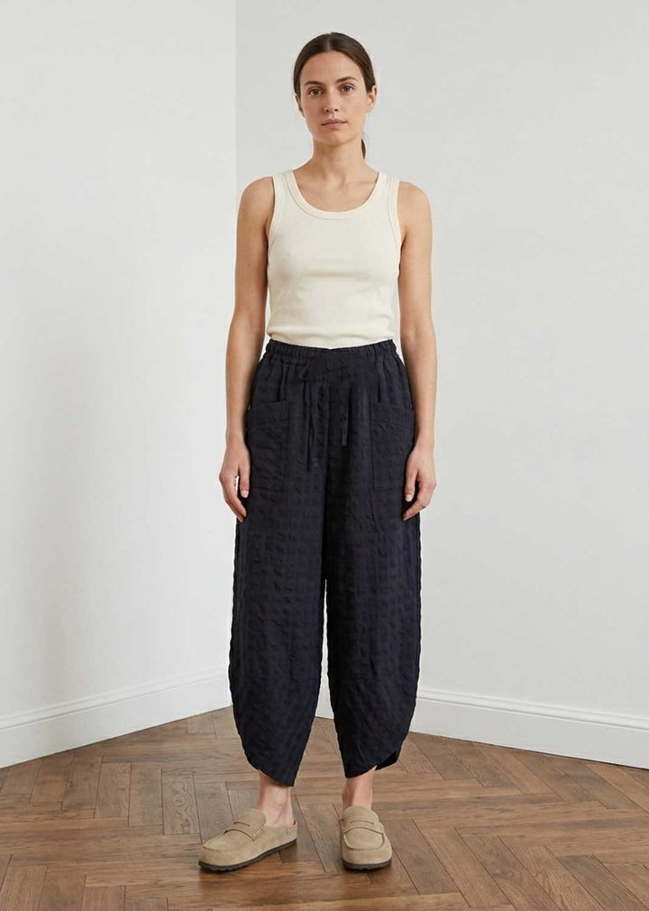A woman stands in a minimalist room with a white wall and wood floor, wearing dark navy textured wide-leg pants, a cream ribbed tank top, and beige slip-on shoes.