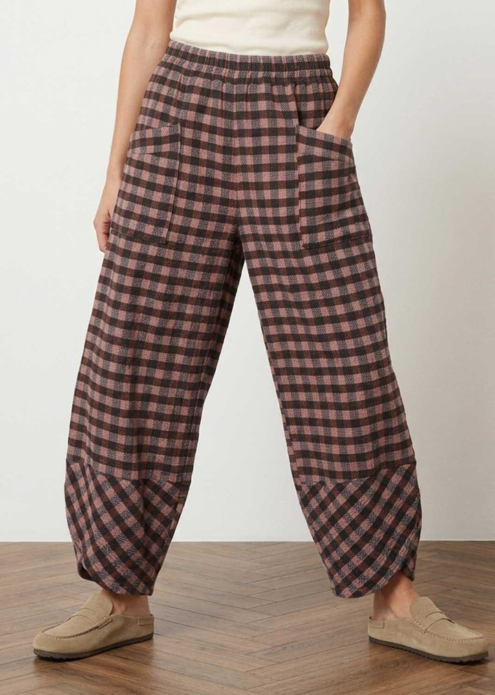 A person wearing loose, wide-leg plaid pants in a pink and dark brown check pattern, paired with a cream top and beige loafers, standing on a wooden floor against a light wall.