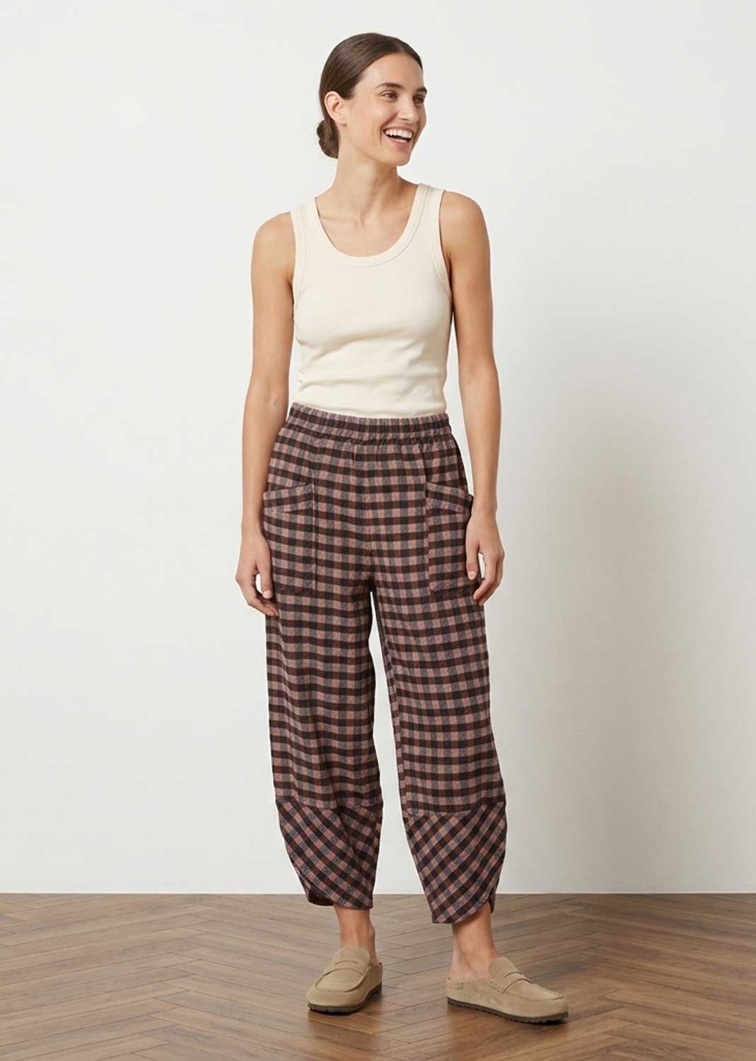 A woman modeling loose, cropped plaid pants in brown and dusky pink tones, paired with a cream tank top and tan slip-on shoes, standing against a plain off-white wall on a wood floor.