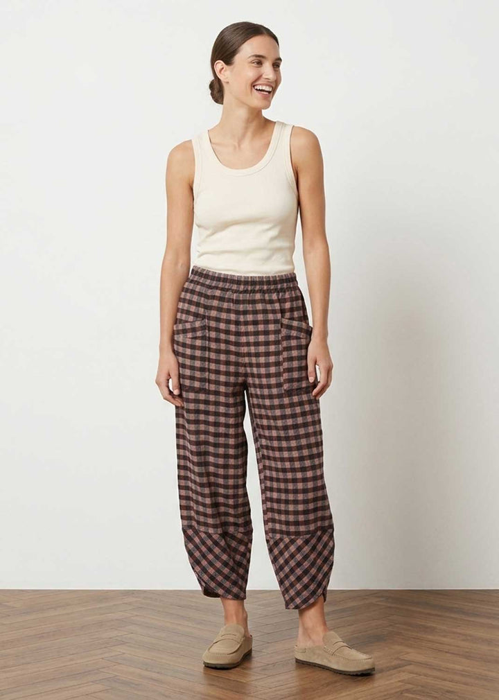 A woman modeling loose, cropped plaid pants in brown and dusky pink tones, paired with a cream tank top and tan slip-on shoes, standing against a plain off-white wall on a wood floor.