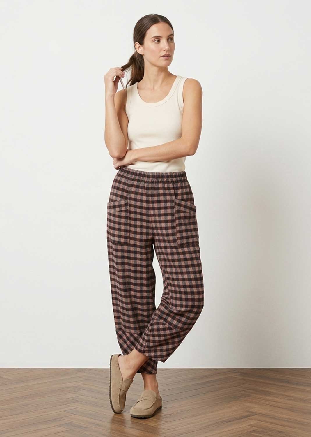 A woman modeling a pair of brown and pink checkered cropped pants with a white tank top and beige slip-on shoes, standing against a white wall on a wooden floor.