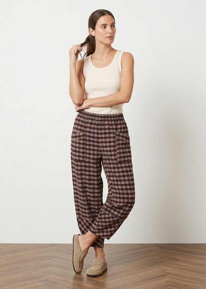 A woman modeling a pair of brown and pink checkered cropped pants with a white tank top and beige slip-on shoes, standing against a white wall on a wooden floor.