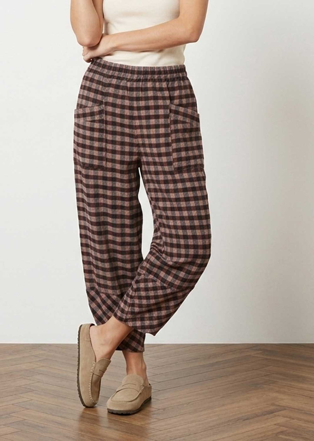 A person wearing loose, high-waisted plaid pants in brown and dusky pink, paired with a cream top and tan slip-on shoes, standing on a wood floor against a light wall.