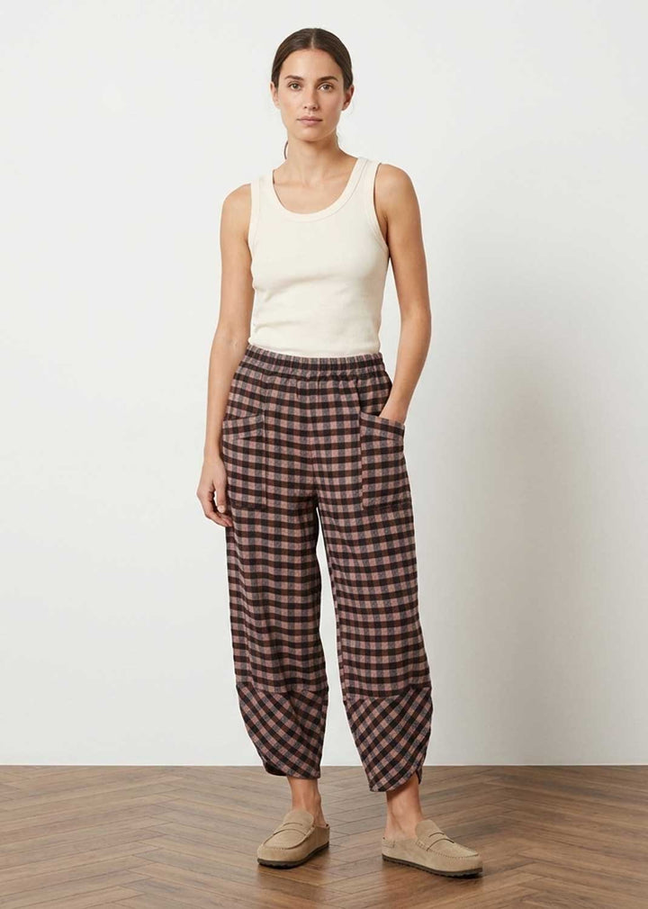 A woman stands in a studio wearing loose brown and pink plaid pants with a cream ribbed tank top and beige slip-on shoes against a white wall and wood floor.