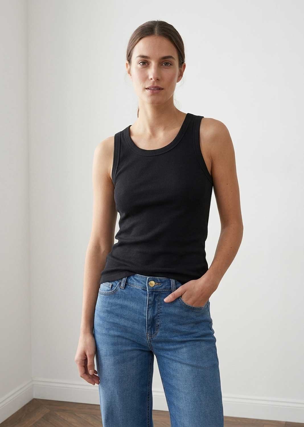 A woman modeling a black sleeveless ribbed tank top paired with blue jeans, standing against a plain white wall.