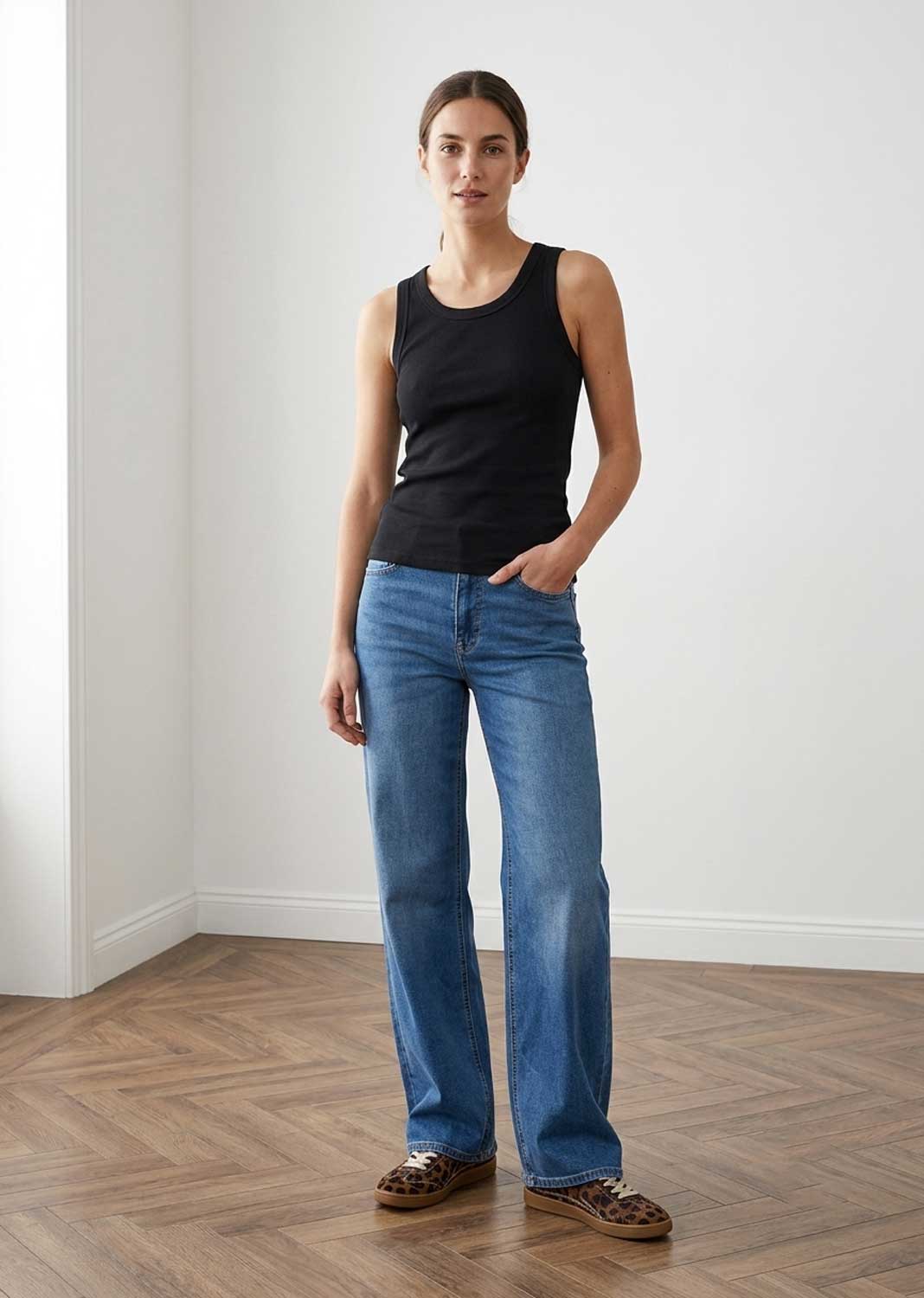 A woman stands in a minimalist room with white walls and a wood herringbone floor, wearing a black sleeveless tank top, blue straight-leg jeans, and brown leopard-print flat shoes.