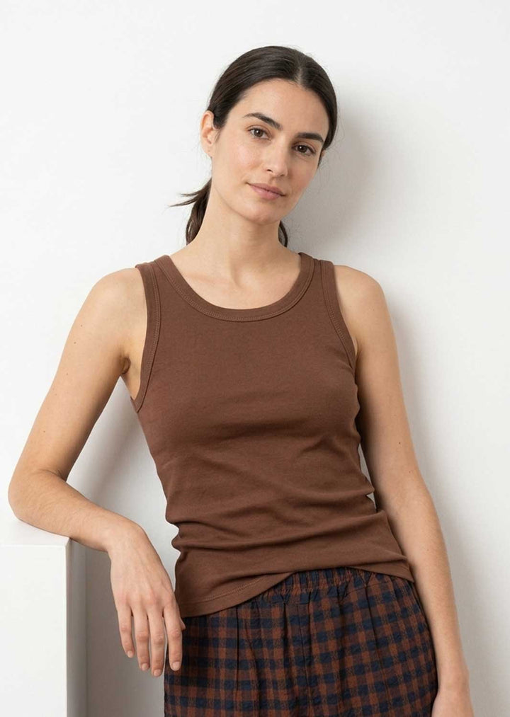 A woman wearing a brown ribbed tank top and a dark plaid skirt, posing against a plain white wall.