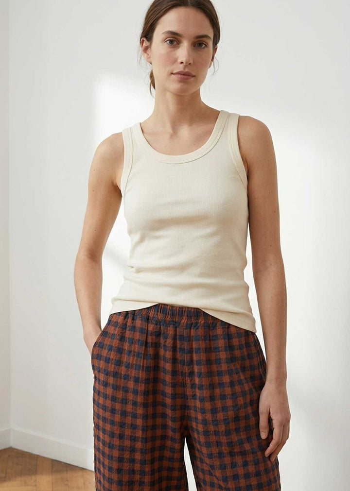 A woman wearing a cream-colored ribbed tank top and dark brown and red plaid pajama pants, standing in a brightly lit room with white walls.