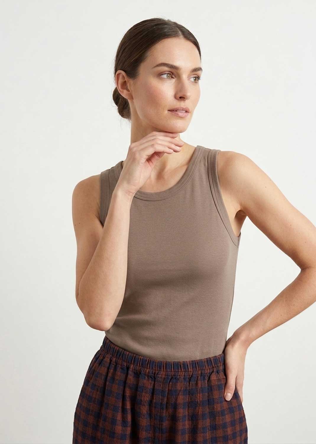 A woman wearing a fitted sleeveless taupe-brown tank top and dark red plaid pants, posed against a clean light gray studio background.
