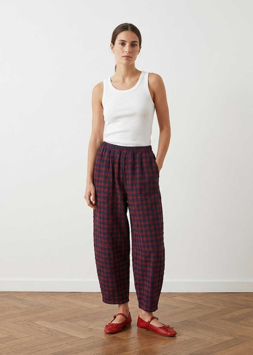 A model stands in a minimalist room with a light gray wall and wooden floor, wearing a white ribbed tank top, dark red and black plaid cropped trousers, and red flat shoes.