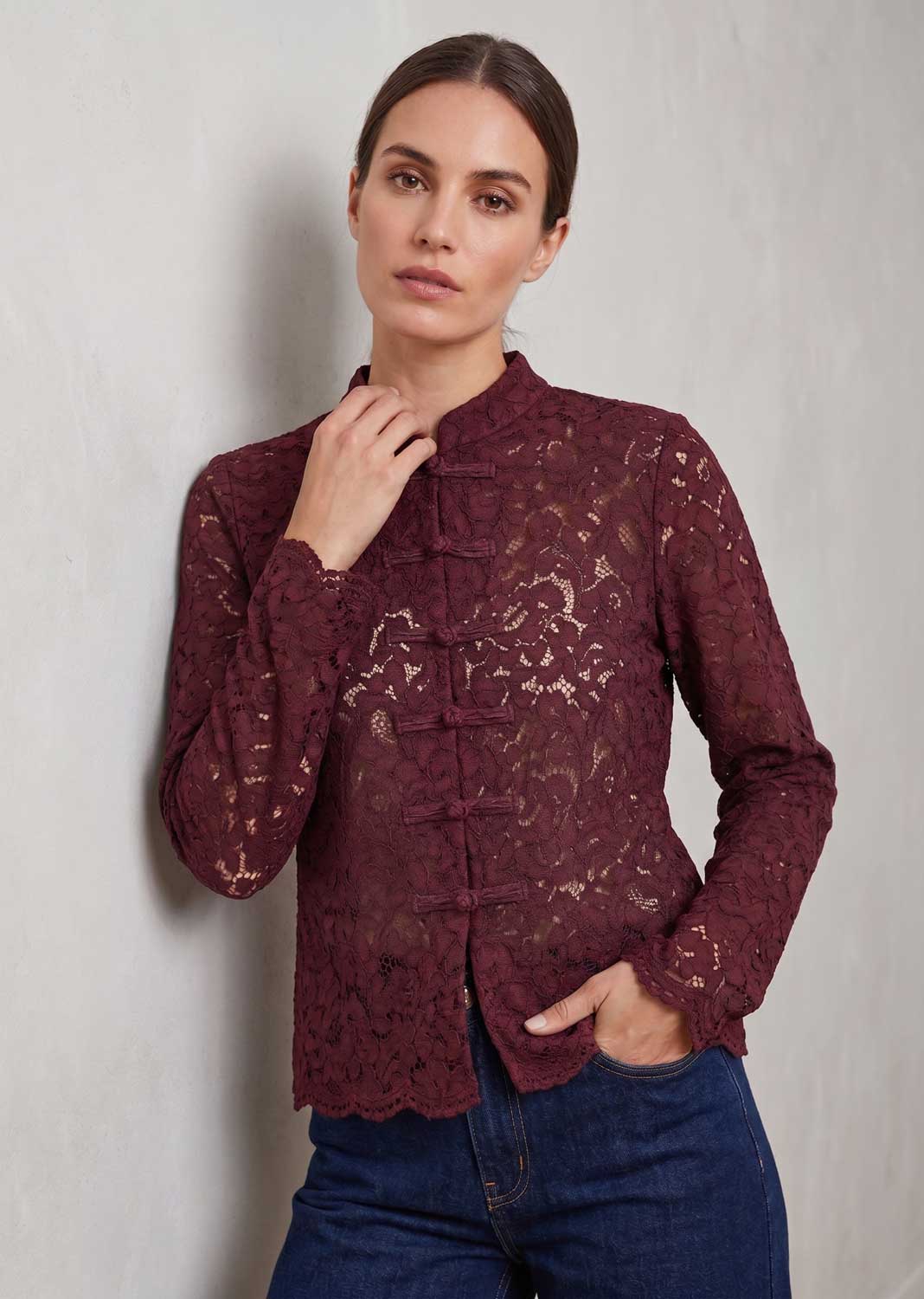 A woman wearing a burgundy lace long-sleeve blouse with a mandarin collar and front button closure, paired with blue jeans, standing against a textured light gray wall.
