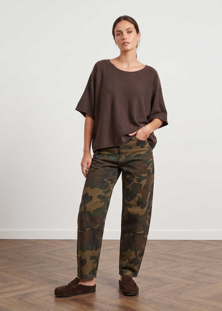 A woman modeling a loose brown short-sleeve top and camouflage print pants, standing on a wooden floor against a plain white wall.