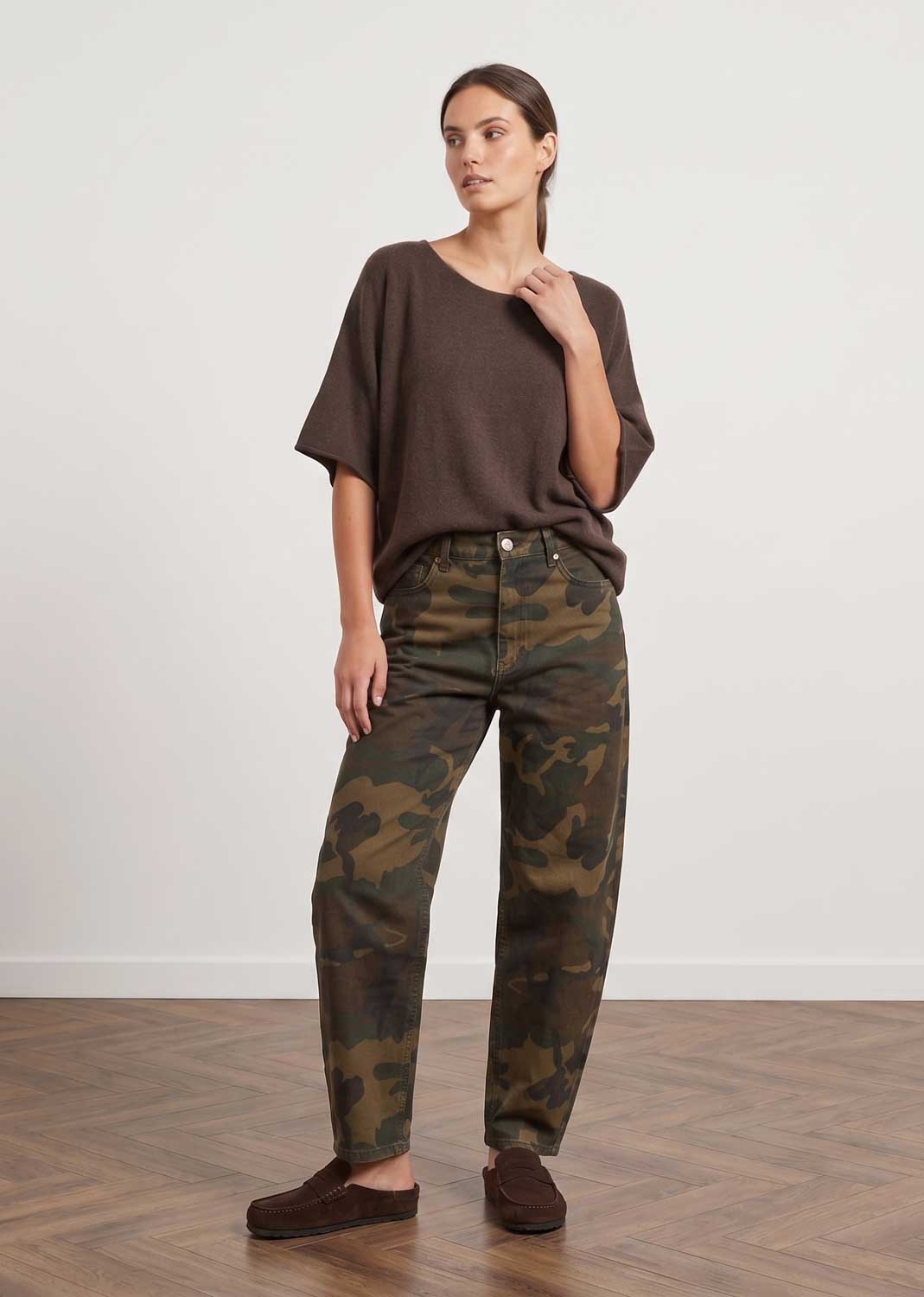A woman modeling a dark brown short-sleeve top and camouflage print pants, standing on a wooden floor against a plain white wall.